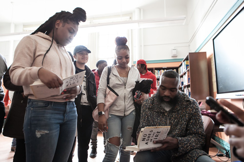 Black man sitting down signing a book. He is surround by standing black and brown students.
