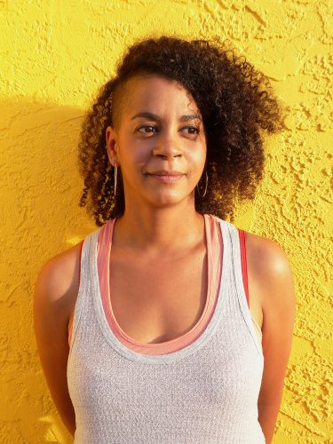 Aisha Sabatini Sloan smiling in front of a bright yellow background. She is wearing a white tank top and large hoop earrings