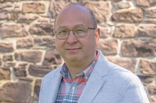 Photo of Jobert E. Abueva giving a small smile in front of a brick wall. He is wearing glasses and a gray jacket over a button up shirt.