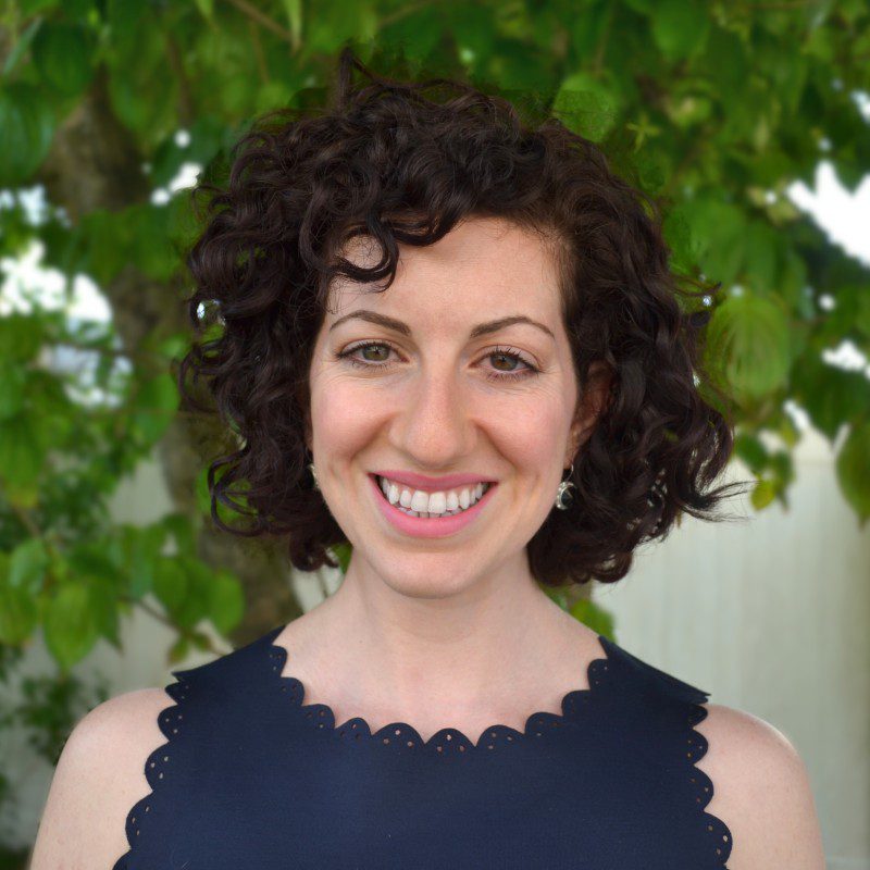 Light-skinned woman with dark curly hair smiling with a tree in the background