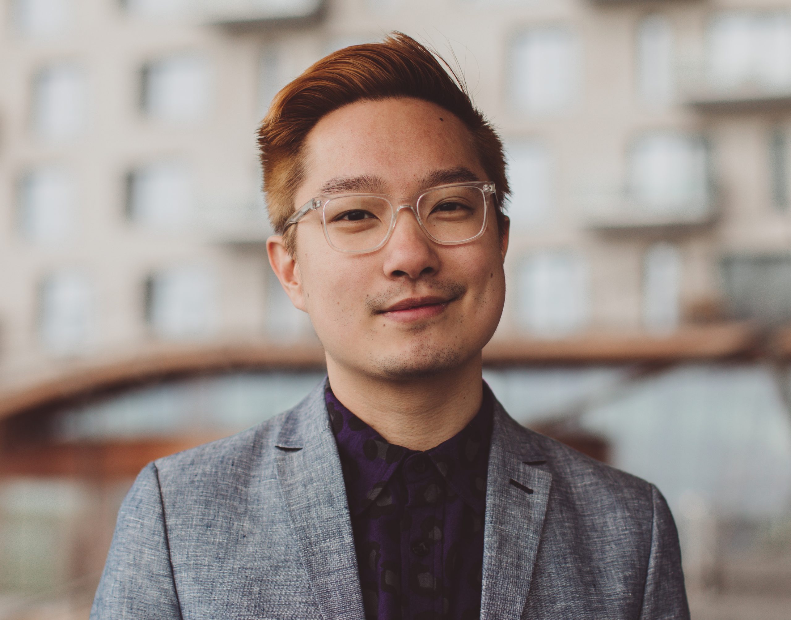 Chen Chen is pictured in front of a blurred building. They have tan-toned skin, short light red hair, and clear glasses framing their face.