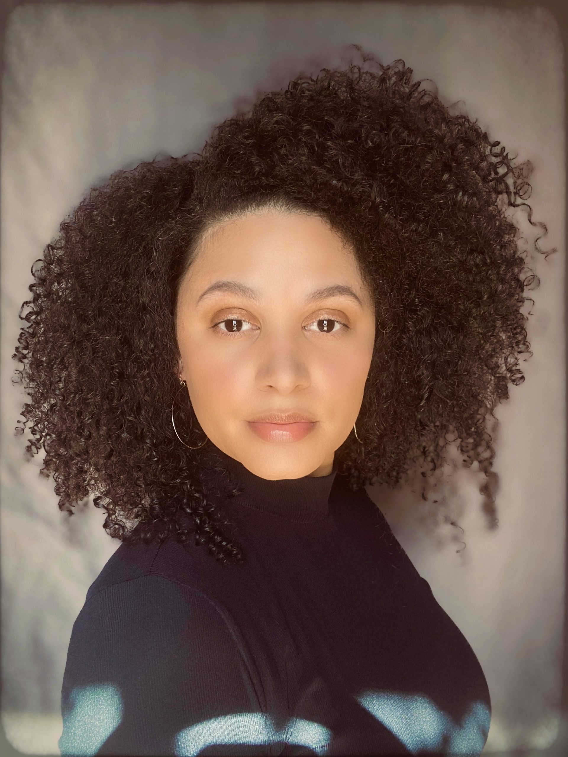 Photographic portrait of author, Kalynn Bayron wearing a black turtleneck against a gray background.