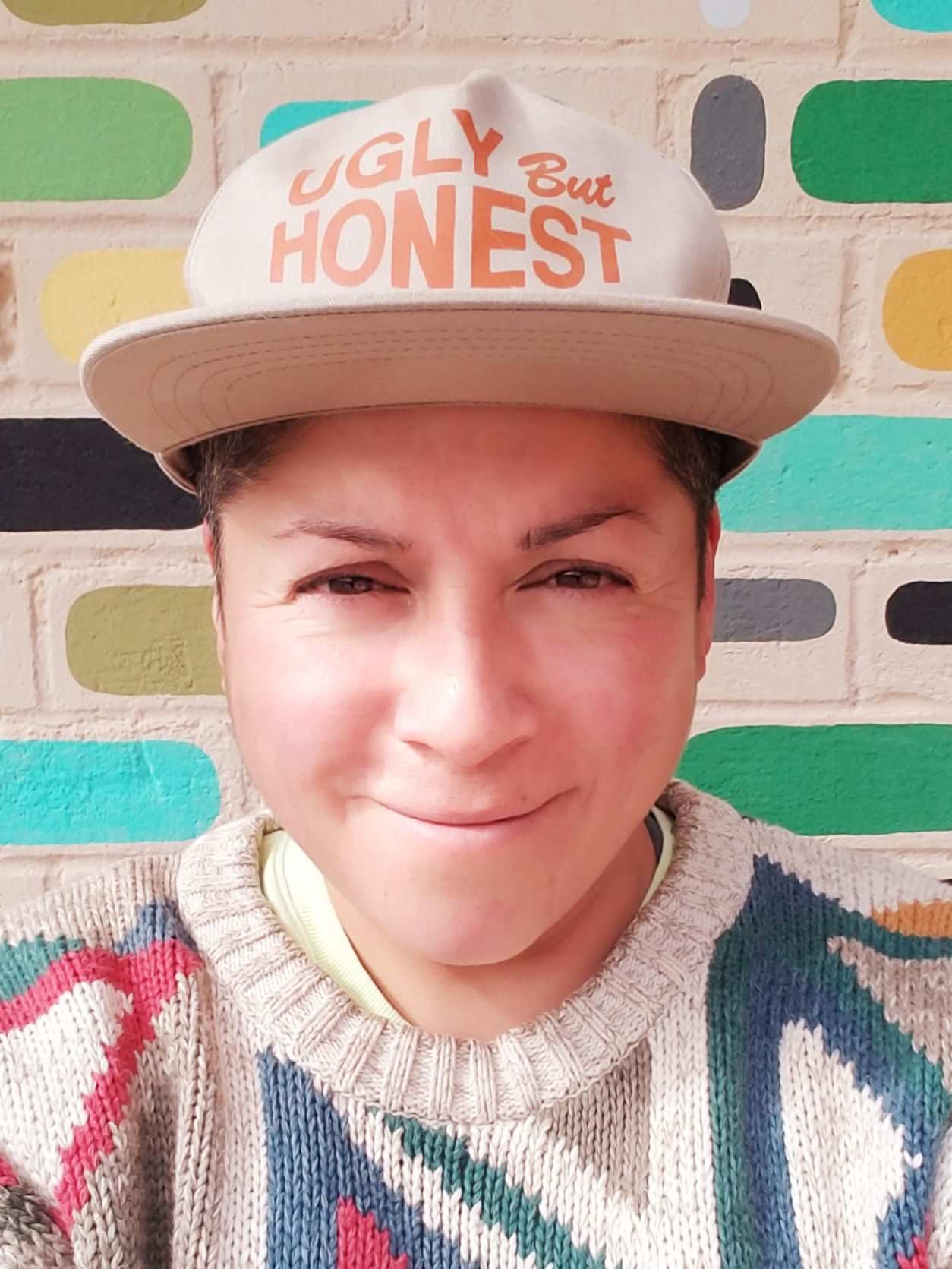 Raquel Gutiérrez is a person with medium skintone and dark hair pictured smiling at the camera, wearing a colorful sweater and a tan hat with the word "Honest" on it.