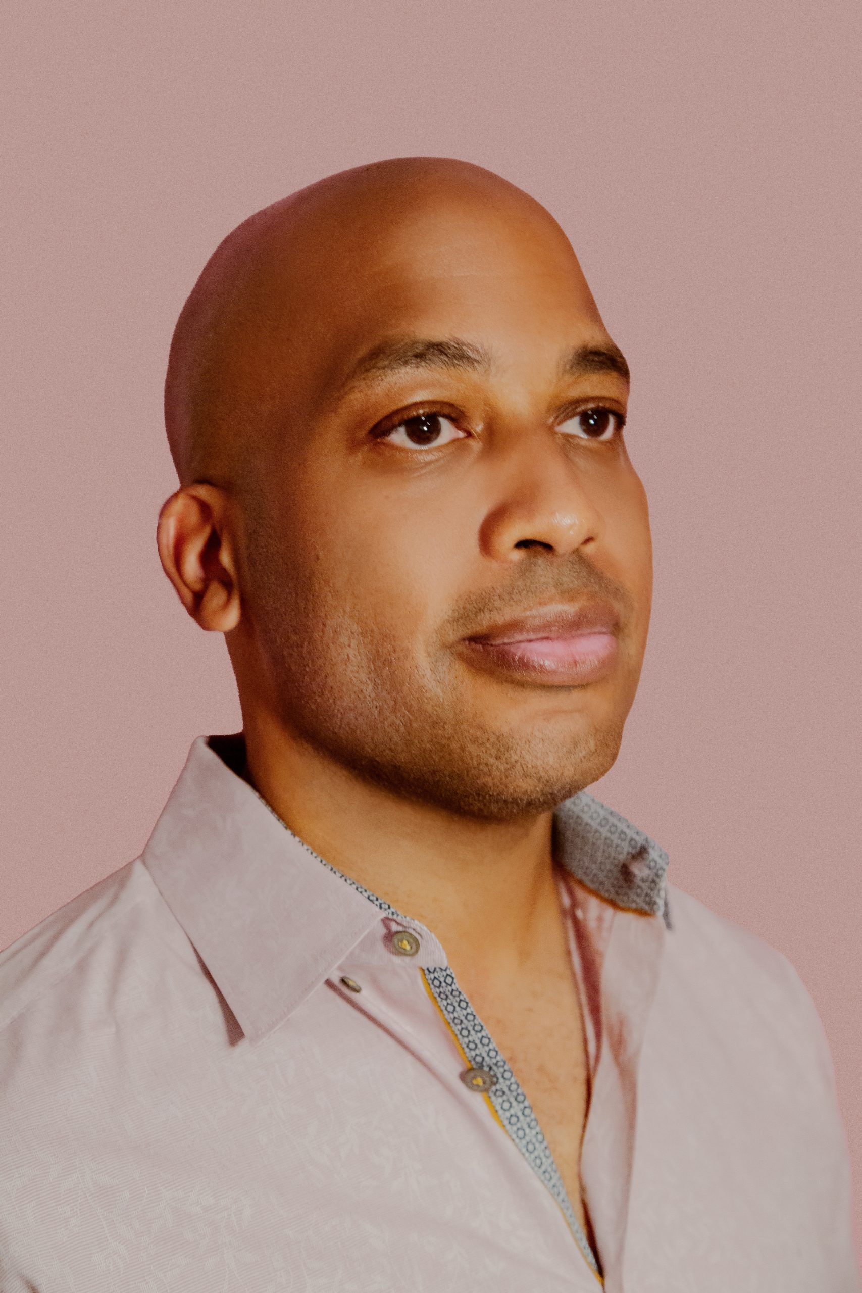 Rasheed Newson is pictured with dark skin tone, a bald head, and a button up shirt, looking off into the distance and smiling slightly
