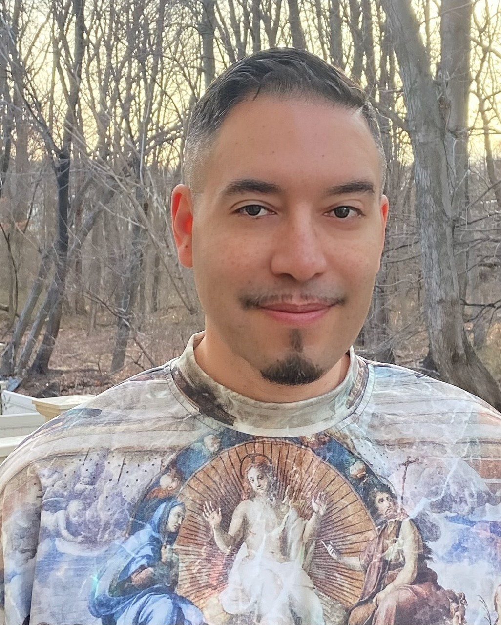 Emanuel Xavier, a Latinx man with light brown skin, short dark hair in a fade cut, and a thin goatee, stands outdoors in a wooded area during late winter. He wears a colorful shirt featuring Renaissance-style religious artwork, and he smiles softly at the camera with bare trees and soft sunlight in the background.