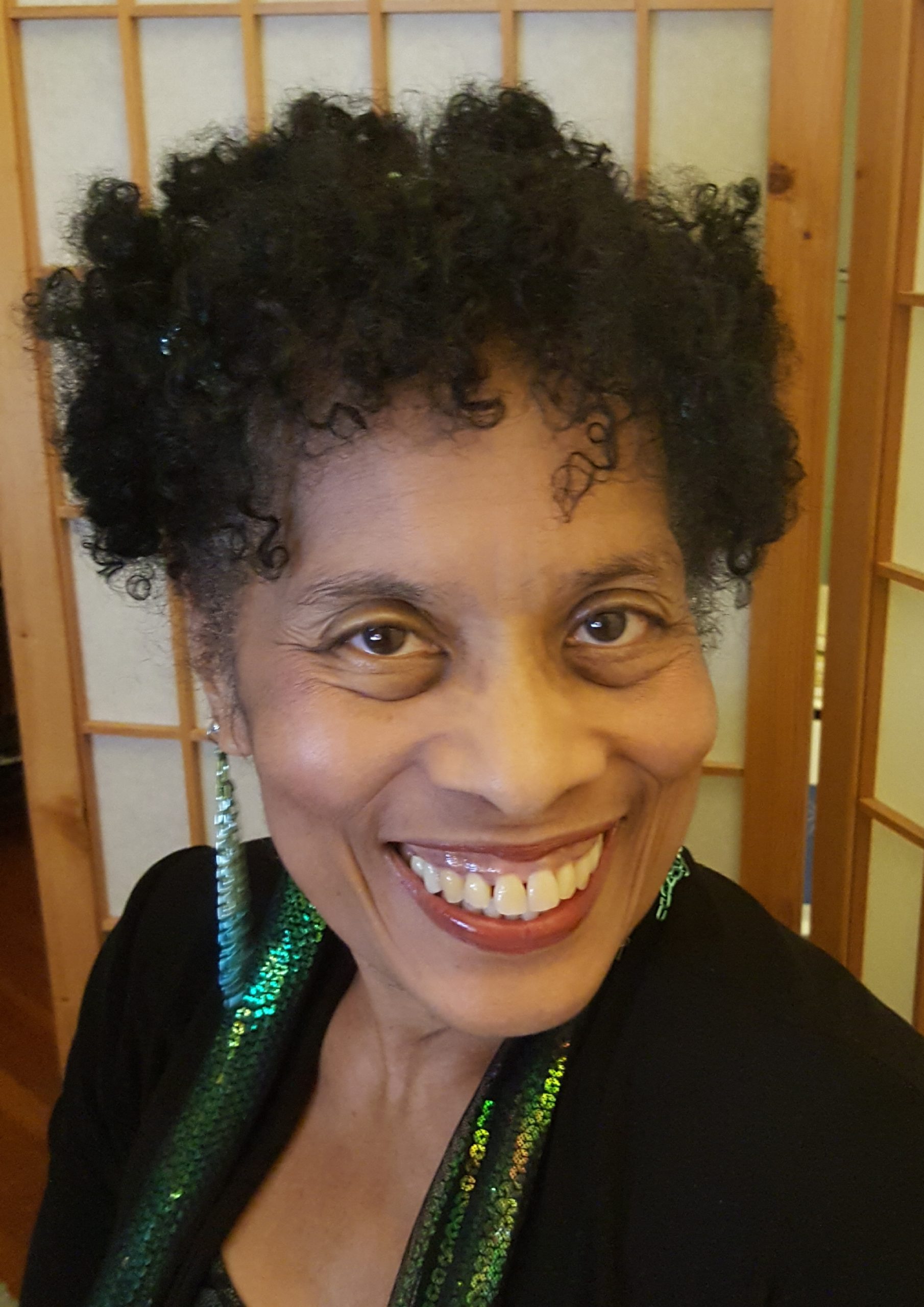 Andrea is a Black woman with curly black hair and a big smile. Her chainmail earrings sparkle.