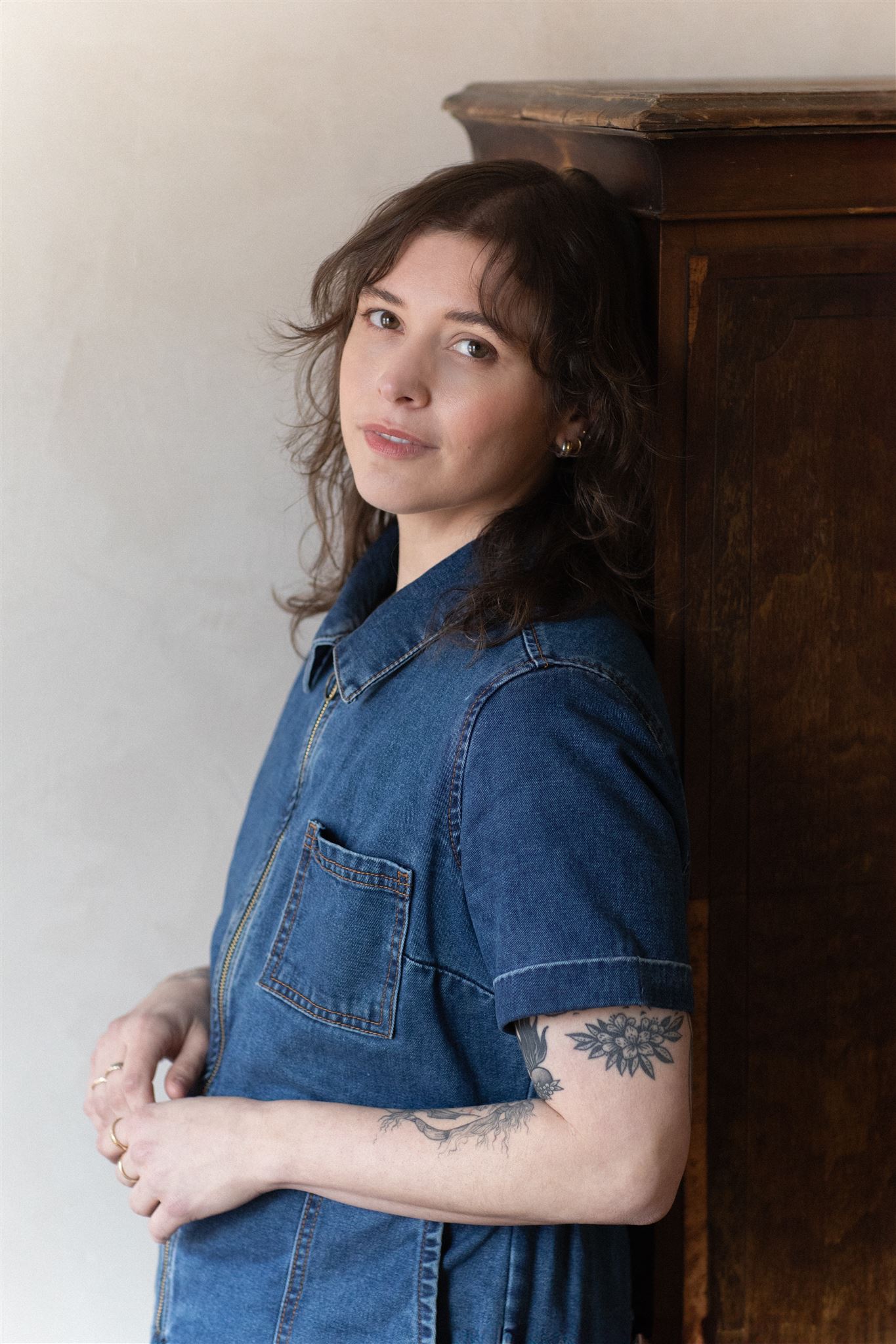 Casey, a white nonbinary person with brown hair and floral tattoos, in a denim jumpsuit