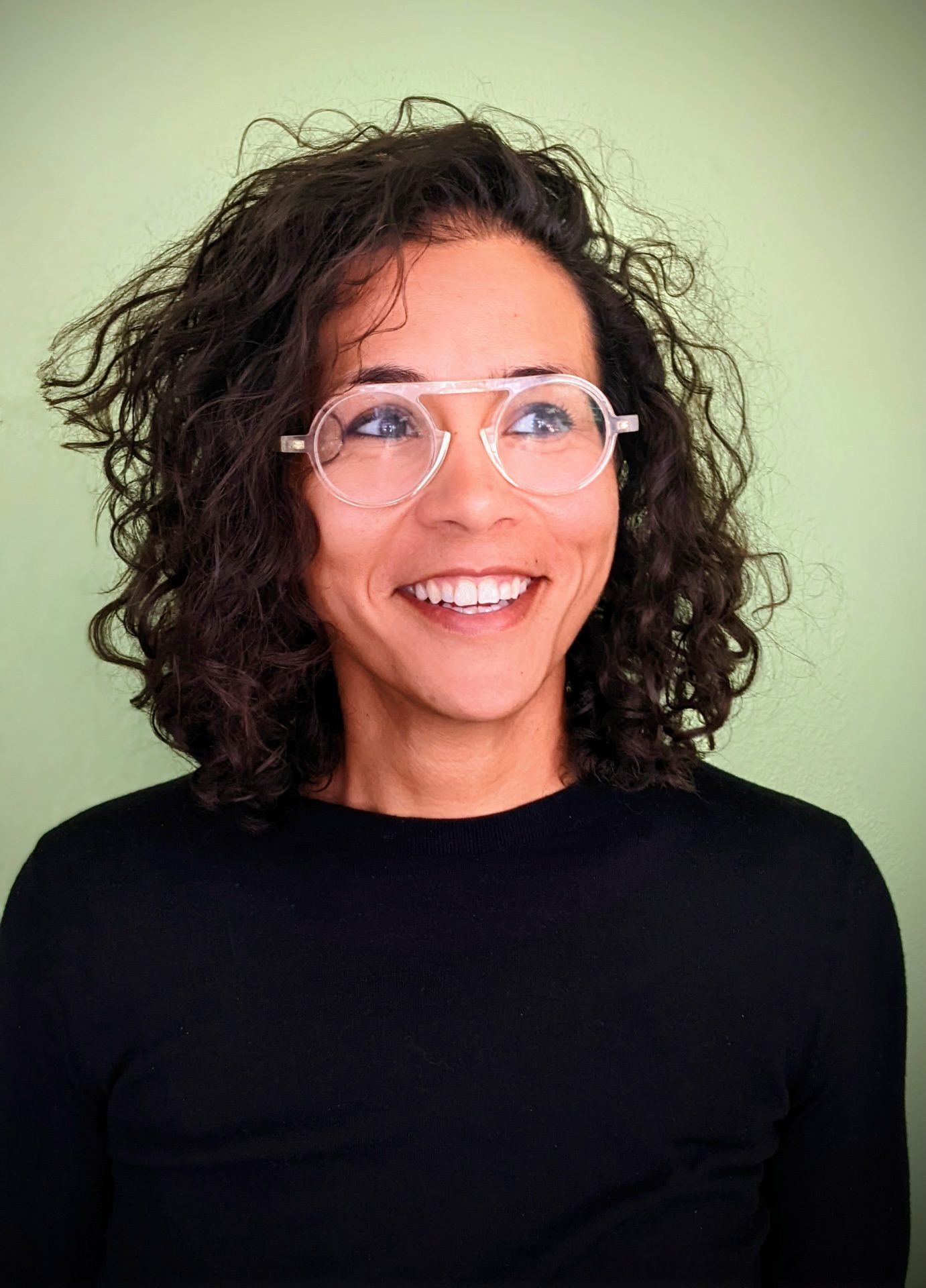 Desta is a biracial woman with curly hair and clear glasses. She is wearing a black shirt against a green background in this semi-formal portrait