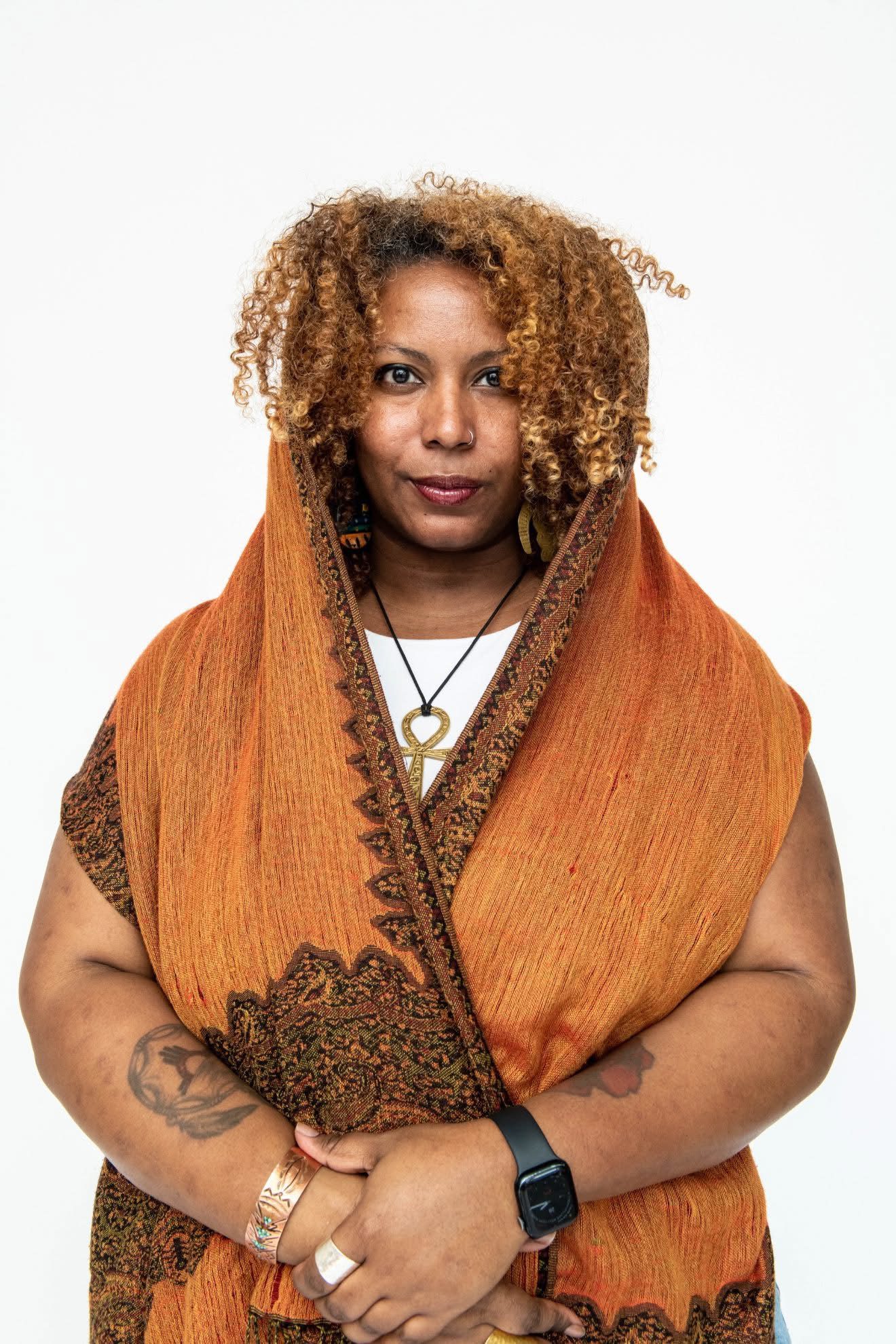An Afro-Latinx person with medium-brown skin and golden-brown curly hair stands against a plain white background. They wear an orange and brown woven shawl draped over their head and shoulders, layered over a white top. They have a small nose ring, visible tattoos on their arms, and wear copper jewelry and a black smartwatch. Their expression is calm and confident.