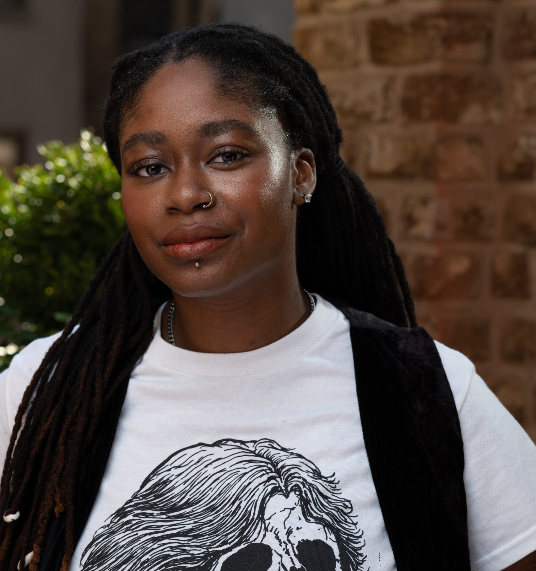 a dark-skinned, Black, AFAB person with long locs in twin ponytails, a labret and a nose piercing. There's a red brick wall behind them and they're wearing a white shirt with a skull on it and a black, velvet vest, with a mischievous grin on their face.