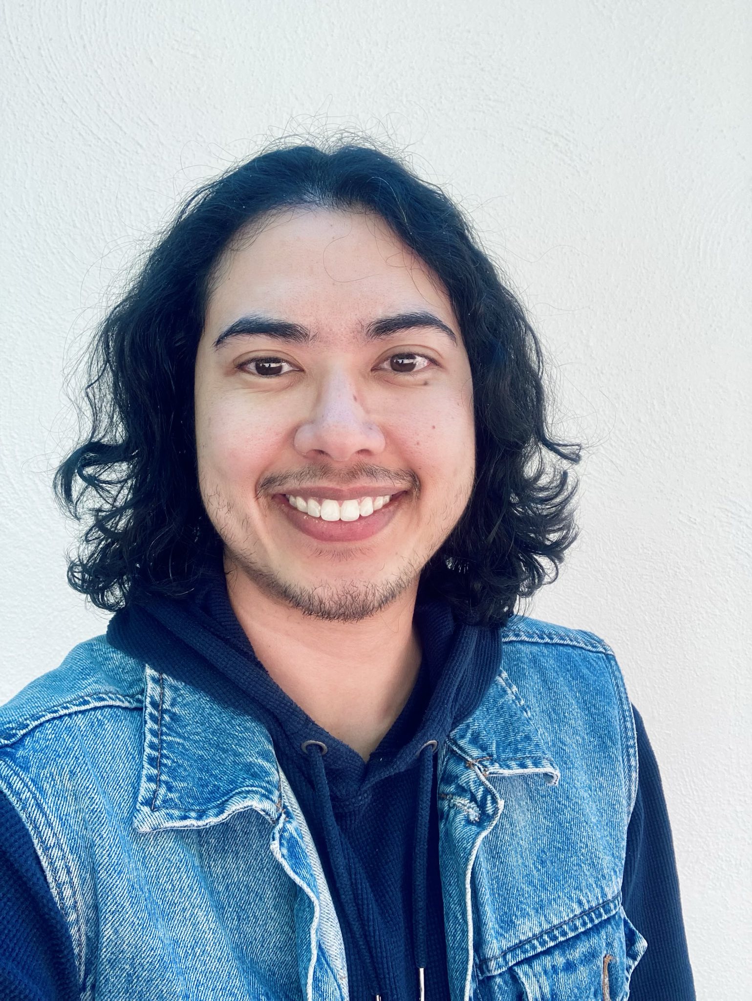 Simo is a nonbinary person of South Asian and Southeast Asian descent with light brown skin, chin-length wavy black hair, dark brown eyes, and some stubble. They are wearing a blue denim vest over a navy blue hoodie and are looking at the camera against a cream-colored background.