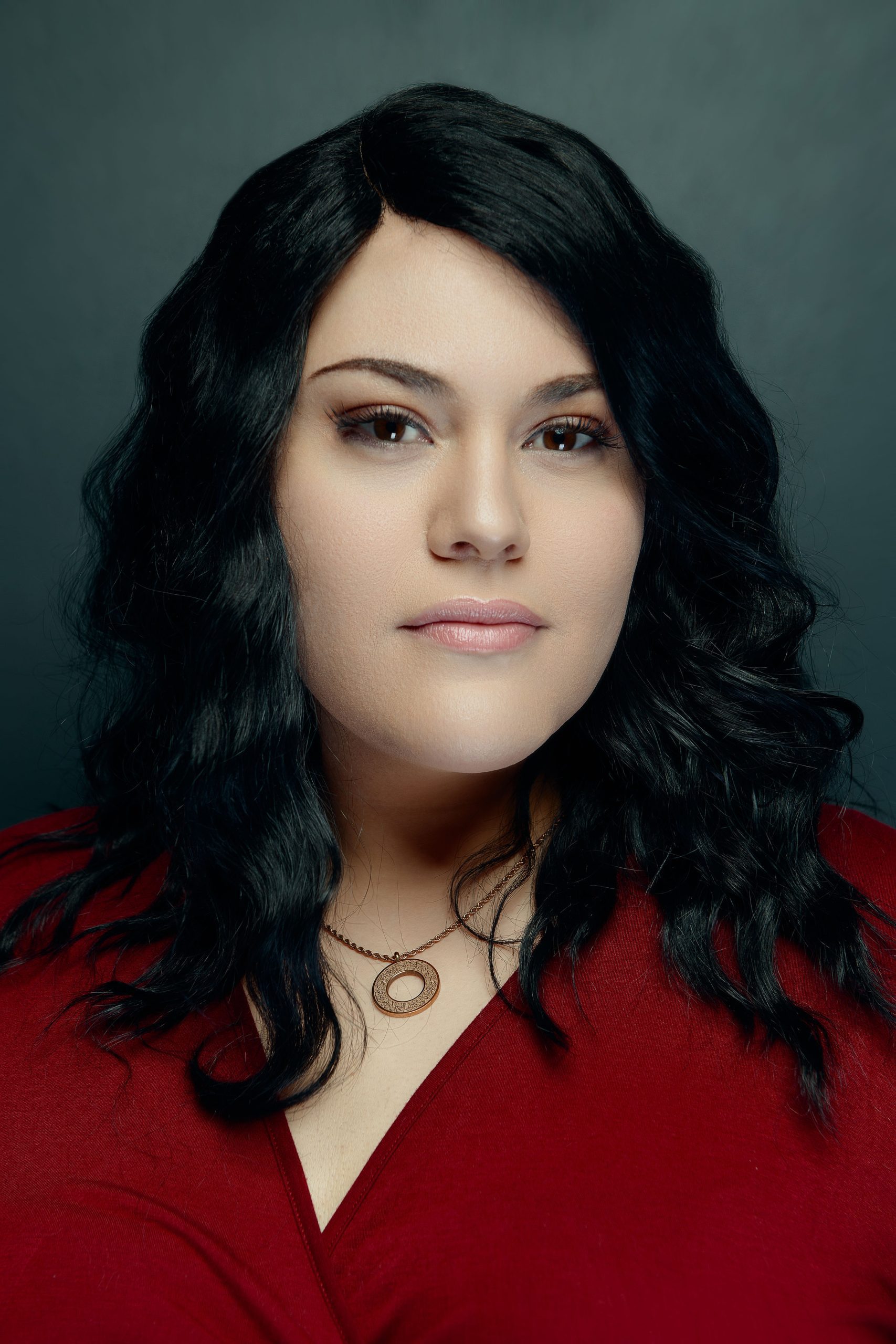 Sharifa is a Arab woman with long wavy black hair. She's wearing a red dress with a rose gold circle necklace in this formal portrait with a dark grey background.