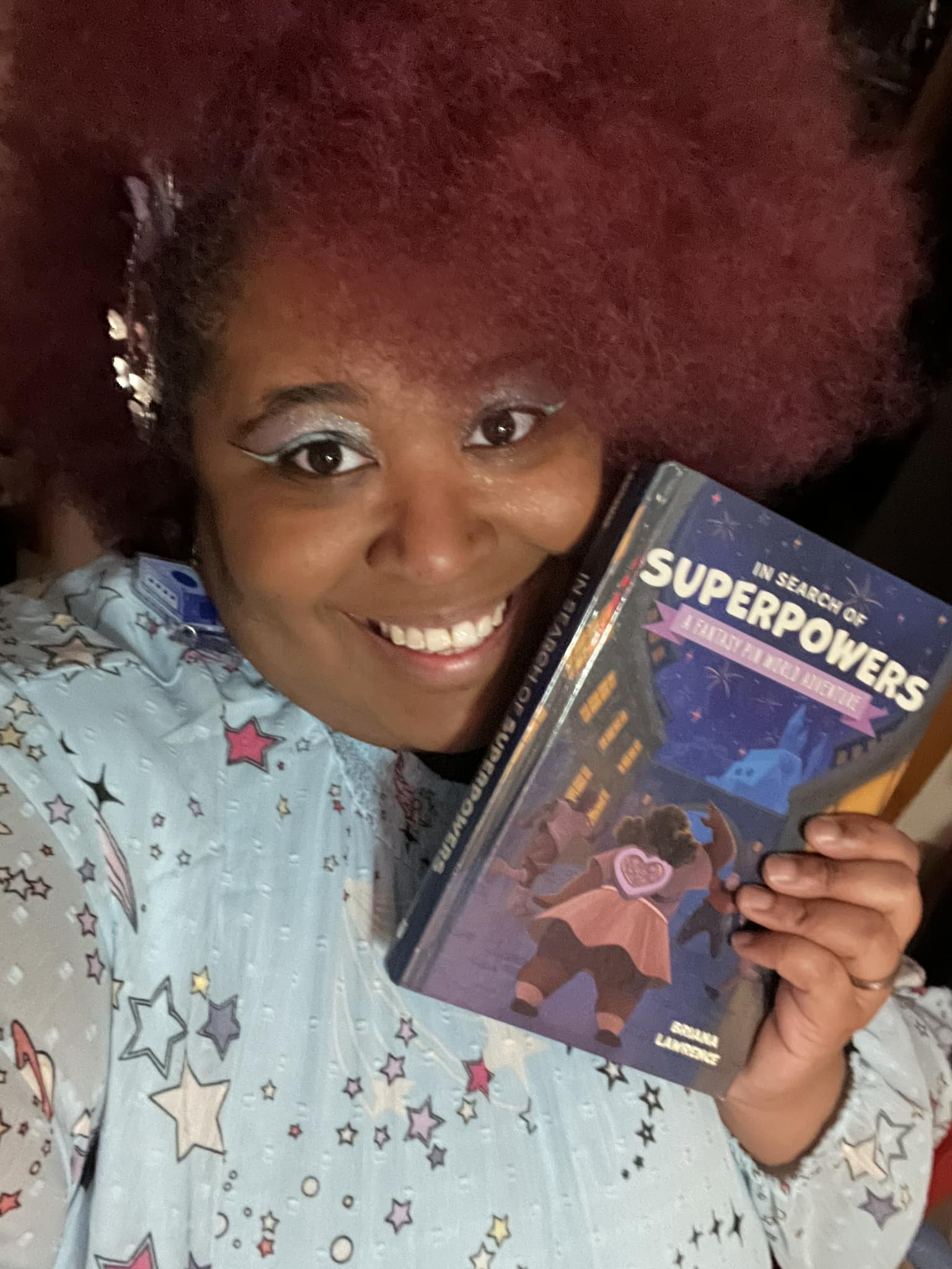 A black woman with reddish, purple hair smiling and holding a hard-covered copy of her book "In Search of Superpowers: A Fantasy Pinworld Adventure." She's wearing a light blue dress that has colorful stars all over it.